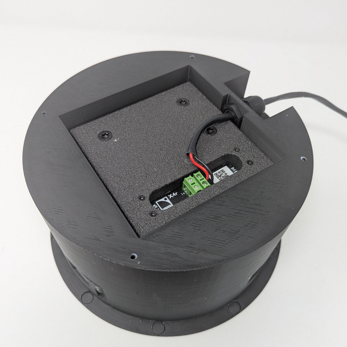 Circular black Terrapin SpkrShell Architectural CW1-X4r In-Ceiling/Wall Enclosure with an open center cavity revealing a terminal block and wiring for X4r in-ceiling installation.